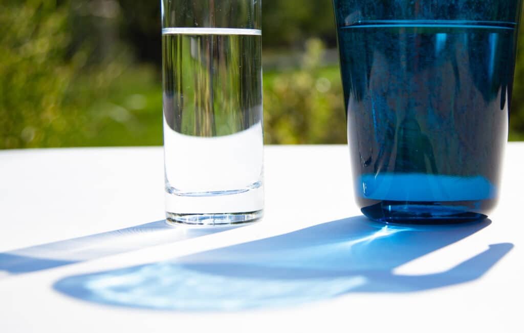 Verre d'eau transparent à côté d'un verre bleu sur une table, avec des ombres et un fond naturel verdoyant.