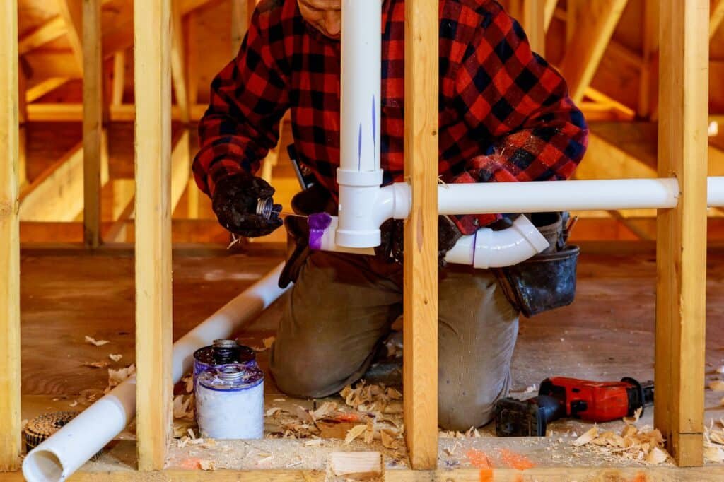 Un artisan en chemise à carreaux travaille sur des tuyaux de plomberie dans une structure en bois, avec des outils et des matériaux de construction autour de lui.