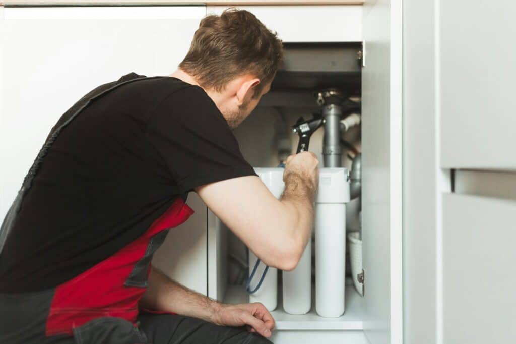 Homme en tenue de travail noire installant un système de filtration d'eau sous un évier dans une cuisine moderne.