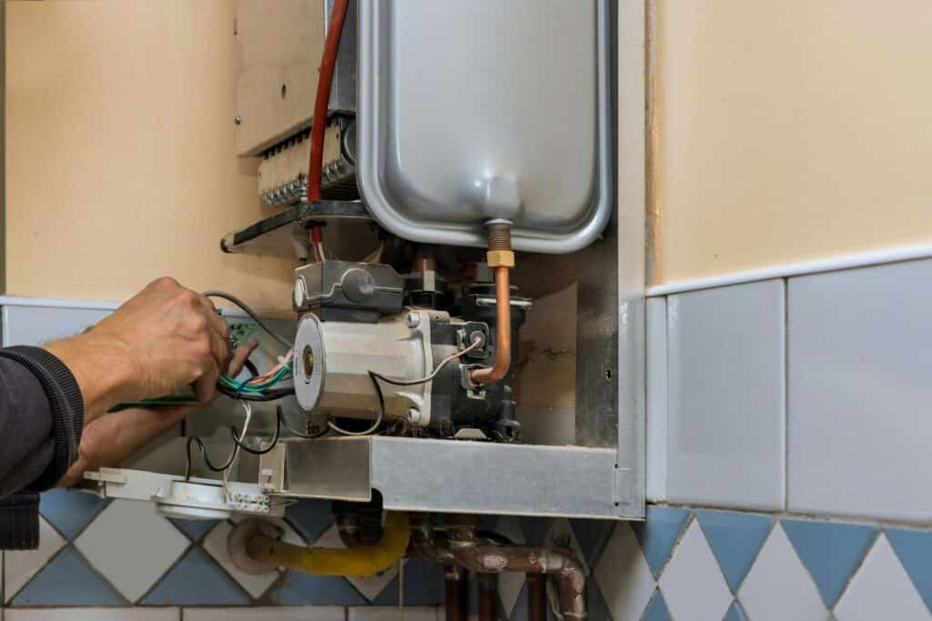 Main d'un technicien ajustant les composants internes d'une chaudière à gaz montée sur un mur de cuisine carrelé.